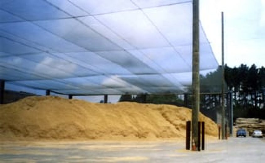 A large pile of wood fiber is covered by a mesh canopy supported by metal poles