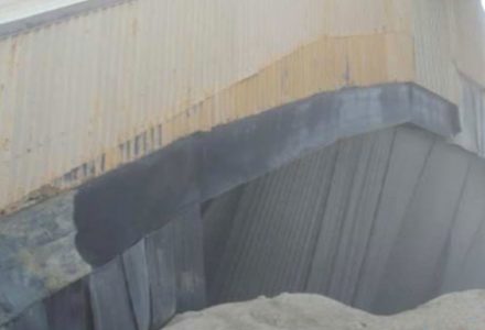 Corrugated metal siding with peeling paint and a dark patch, above a pile of light-colored material