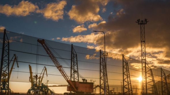 A construction site with cranes and a large wind fence at sunset