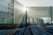 Railway tracks curve gently beside tall, translucent wind fences, illuminated by sunlight