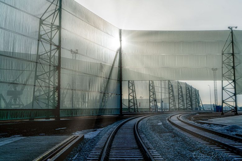 Railway tracks curve beside tall, translucent wind fences with sunlight filtering through