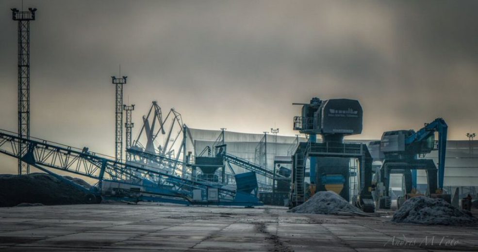 Industrial site with conveyor belts, cranes, and piles of material under a cloudy sky