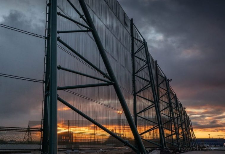 A large industrial fence structure with a sunset visible through its mesh panels