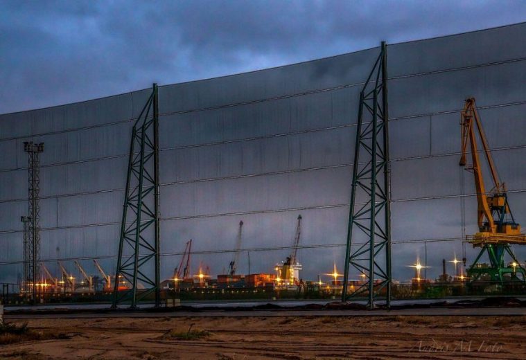 A large industrial wind fence stands in front of a port with cranes and ships