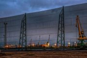 A large industrial wind fence stands in front of a port with cranes and ships