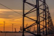 A metal structure stands tall against a vibrant sunset sky with cranes in the background