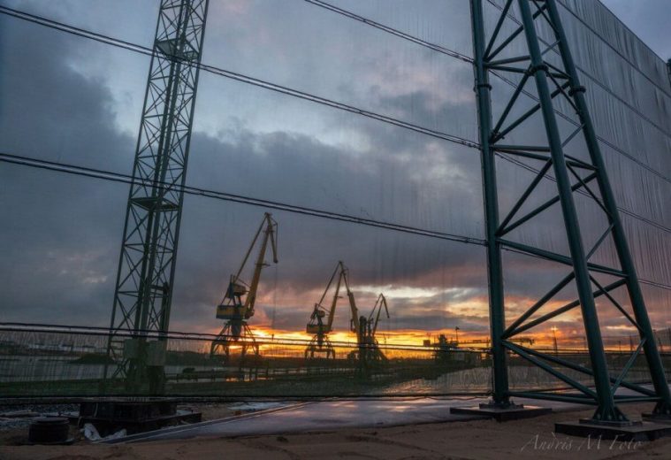 Cranes silhouetted against a sunset, viewed through a metal framework and netting