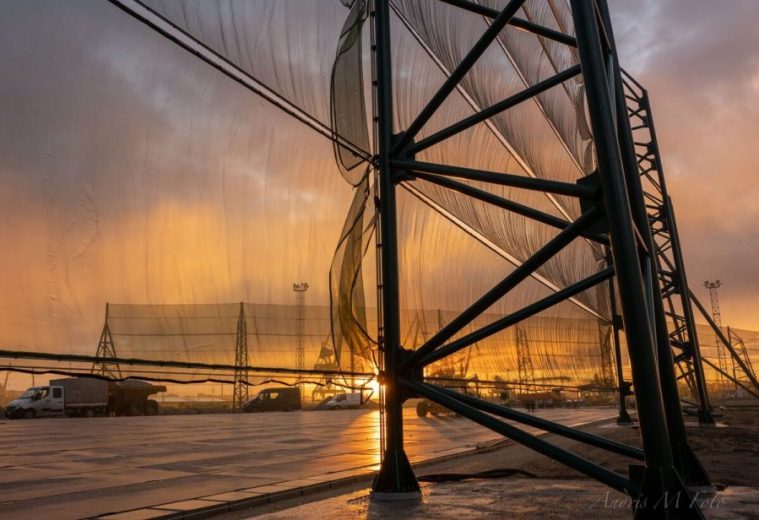A large wind fence structure stands against a dramatic sunset sky