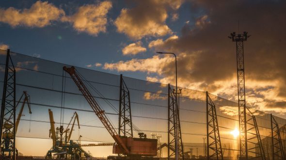 A crane and industrial equipment are visible behind a large wind fence at sunset