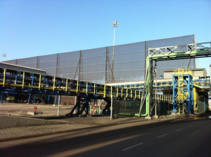 An industrial conveyor belt system with yellow and blue supports and a large building in the background