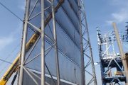 A tall metal windbreak structure stands against a clear blue sky with construction equipment nearby