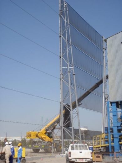 A tall industrial wind fence with workers and machinery at the base under a clear sky
